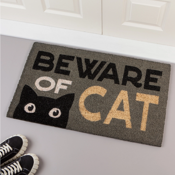 A grey doormat featuring the words "Beware Of The Cat" in bold, with a cartoonish black cat peeking over the edge.