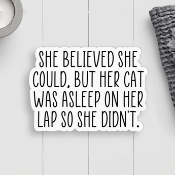 A white sticker with black text that reads "She Believed She Could, But Her Cat Was Asleep On Her Lap So She Didn't," featuring a cozy cat illustration.