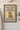 Framed cross-stitch art with a cat and text 'Welcome to the Bathroom, a cat will be with you shortly' on a tiled bathroom wall.