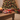 A festive red ceramic mug with Christmas-themed illustrations of a snowman, Christmas tree, Santa, and a star, resting on a wooden table in a cozy holiday setting with a blurred Christmas tree in the background.