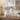 A white ceramic cat figurine with black stripes on its back, styled on a wooden table next to a gold lamp, a white candle, and a small succulent in a blurred living room background.