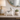 A glossy white ceramic cat figurine with black stripes and facial details sits on a light stone console table in a styled interior setting, alongside a lamp, a small succulent, a candle, and a round mirror.