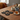 Four black cat-shaped coasters with embroidered yellow, white, and pink details are arranged on a wooden table in a cozy indoor setting, surrounded by mugs of tea and coffee, candles, and a knit blanket.