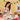 A young girl wearing a festive Christmas apron sits at a table decorated for the holidays, mixing ingredients in a bowl. The scene features matching Christmas-themed tableware, including bowls, mugs, and cookie cutters, along with a decorated Christmas tree and a yellow wall in the background.