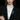 A close-up of a model wearing a black blazer over a white blouse with a large bow, featuring a colorful cat-shaped brooch pinned to the left lapel. The background is solid black.