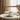 A white ceramic cat-shaped candle holder with gold wire glasses and a gold tail, sitting on a matching white saucer on a round white side table in a softly lit room.