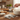 A hand uses a gray measuring scoop with a cat face to pour flour into a white bowl on a wooden table, with matching gray measuring cups and spoons, glass jars, a rolling pin, and a whisk in a warm kitchen setting.