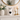 A white ceramic cat figurine with black glasses and a black heart on its chest, displayed on a white mantelpiece alongside a white ribbed vase with flowers, two lit white pillar candles in gold holders, and a round gold-framed mirror.