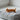 A small orange cat figurine with orange stripes stands on top of a light gray quilted zippered pouch on a white surface near a window.