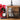 A pair of gray Christmas-themed socks with black cats wearing red Santa hats and the text 'MEOW KIT CHRISTMAS' hangs from the front of a rustic wooden chest of drawers. The scene is styled with a red poinsettia, a small black-and-white clock on stacked books, and a black table lamp against a white wood-paneled wall.