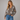 A woman wearing a brown knit cardigan with a cat pattern, paired with blue jeans, against a solid gray studio background.