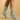 A pair of ankle socks worn on a model's feet, featuring a light mint green background with a whimsical print of cacti, boots, and a cat. The background is a solid, warm beige studio backdrop.