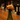 A small orange cat-shaped bottle stopper with closed eyes and pink stripes rests on the neck of a green wine bottle on a wooden bar counter, with a blurred bar background.