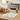 A white fabric pet care mat with a colorful cartoon cat pattern is laid out on a wooden kitchen table next to a small blue pill organizer and an amber medicine bottle. In the background, a wooden cutting board holds fresh apples, carrots, blueberries, and parsley, with a blurred kitchen setting behind.