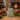 A light sage green ceramic cat figurine sits on a wooden surface surrounded by pine branches, red berries, pinecones, and warm fairy lights, with a blurred Christmas tree and fireplace in the background.